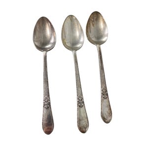 May include: Three silver-toned metal spoons arranged vertically. Each spoon features an oval bowl and a slender handle decorated with a raised floral motif near the base, characterized by a metallic sheen and visible signs of aging.