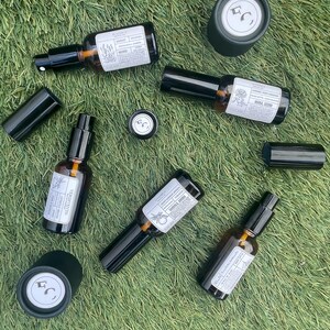 May include: A collection of five amber glass spray bottles with black caps and white labels, accompanied by two separate black cylindrical caps. Each label features printed text and botanical illustrations, with some bottles showing the brand logo.