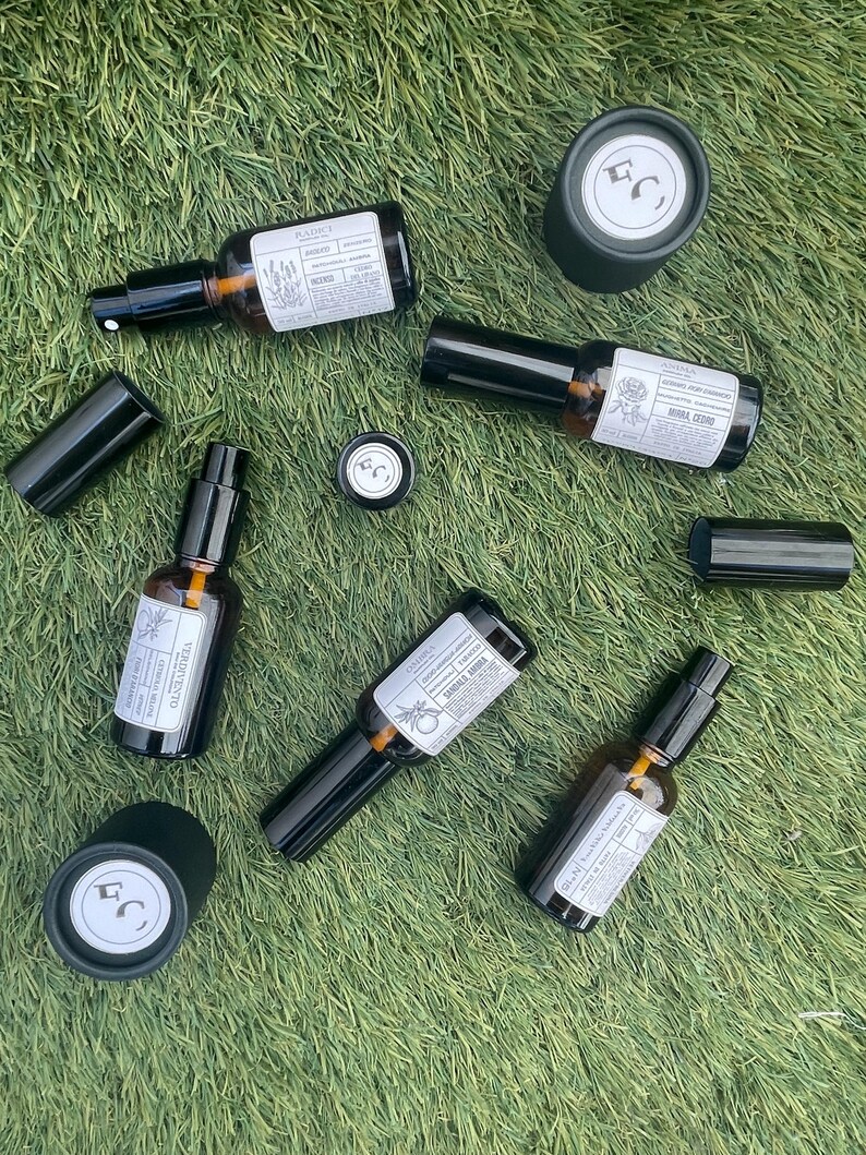 May include: A collection of five amber glass spray bottles with black caps and white labels, accompanied by two separate black cylindrical caps. Each label features printed text and botanical illustrations, with some bottles showing the brand logo.