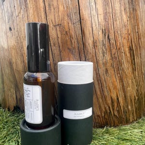 May include: A brown glass spray bottle of Radici parfum oil with a black cap and a white label featuring text, standing next to a black cylindrical cardboard container with a white lid and a white label reading Radici Eau de Parfum.