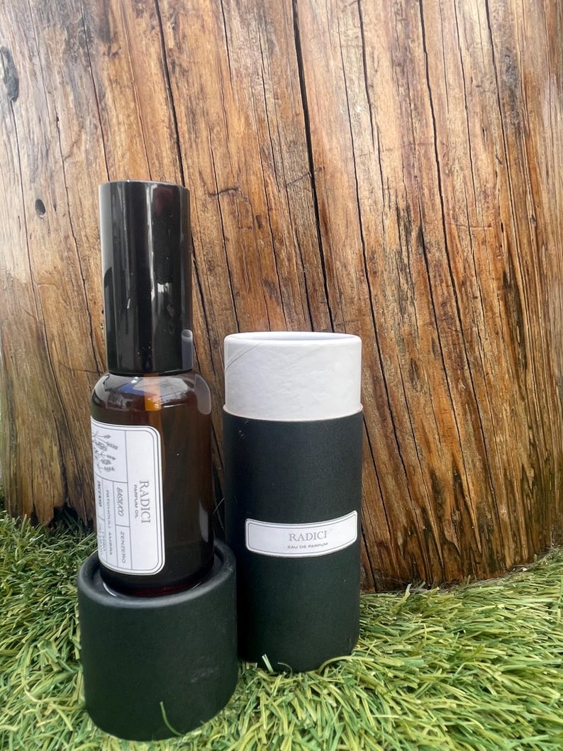 May include: A brown glass spray bottle of Radici parfum oil with a black cap and a white label featuring text, standing next to a black cylindrical cardboard container with a white lid and a white label reading Radici Eau de Parfum.