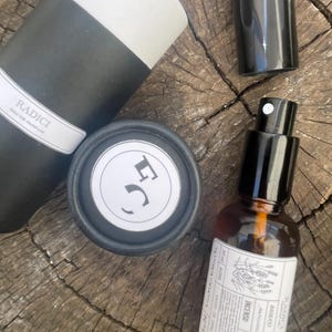 May include: A Radici Eau de Parfum set including a black and white cylindrical container, a small black cap, and an amber glass spray bottle with a white label featuring the text Radici, Patchouli, Ambra, Cedro, Incenso, and 30 ml.