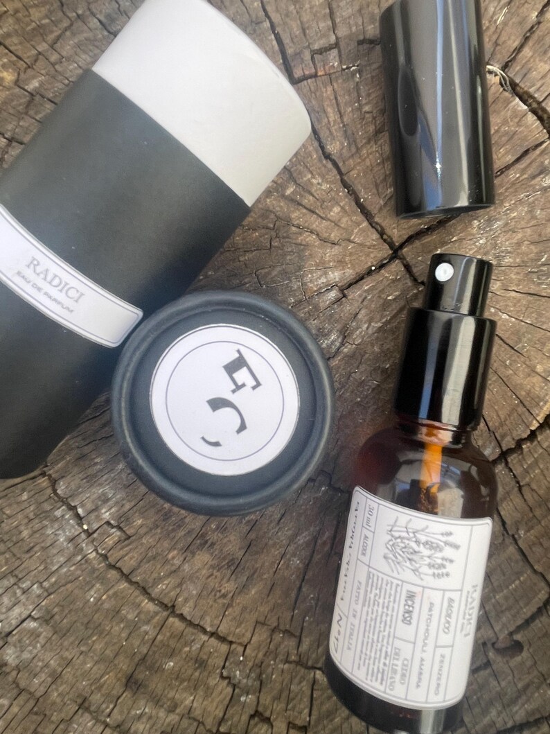 May include: A Radici Eau de Parfum set including a black and white cylindrical container, a small black cap, and an amber glass spray bottle with a white label featuring the text Radici, Patchouli, Ambra, Cedro, Incenso, and 30 ml.