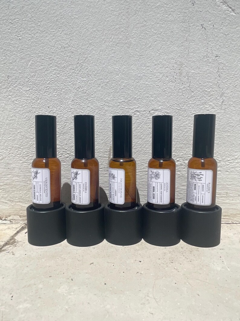 May include: Five amber glass spray bottles with black caps, each held in a cylindrical black base. The bottles feature white rectangular labels with black text and botanical illustrations, displaying the names Verivento, Ombra, Nightborne, Anima, and Radici.