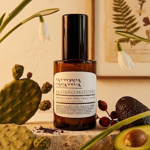 May include: A brown glass bottle of Fattoria Creativa Blossomousse face cream with a black cap. The white label features black text including the product name and ingredients. The bottle is surrounded by cactus pads, snowdrop flowers, and an avocado.