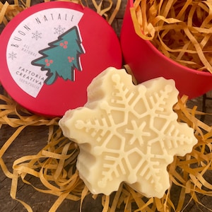 May include: A snowflake-shaped, off-white soap bar rests on shredded paper, near a red container and a red lid with text and a Christmas tree design. The lid reads "BUON NATALE" and "ALL NATURAL".