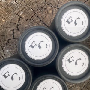 May include: Five cylindrical black containers arranged in a cluster. Each container features a circular white label on the top lid, printed with a black logo consisting of the letters GC in a stylized font. The containers have a matte finish.