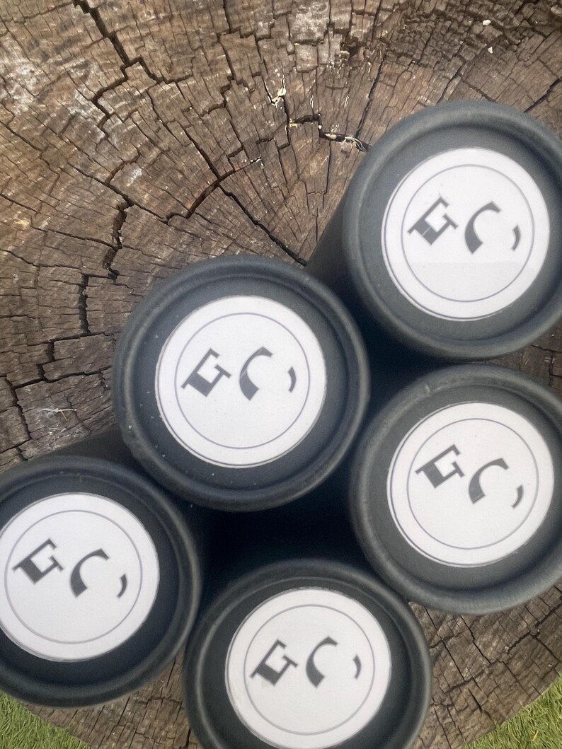 May include: Five cylindrical black containers arranged in a cluster. Each container features a circular white label on the top lid, printed with a black logo consisting of the letters GC in a stylized font. The containers have a matte finish.