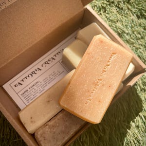 Può includere: Una scatola con cinque saponi profumati diversi. I saponi sono rettangolari e di colore marrone chiaro. La scatola è di cartone e di colore marrone. Il testo "FATTORIA CREATIVA" è stampato sulla scatola. Il testo "Prodotti di Saponeria" è stampato su uno dei saponi.