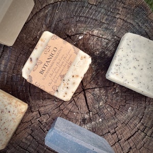 Può includere: Quattro saponette fatte a mano su una superficie di legno. Le saponette sono etichettate "BOTANICO (LAVANDA&ROSMARINO)" e elencano gli ingredienti, tra cui olio d'oliva, olio di cocco, rosmarino, lavanda, olio di ricino, burro di karitè e olio di jojoba.