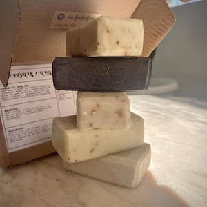 Può includere: Una pila di quattro saponette rettangolari, con una scatola di cartone marrone dietro. Le saponette sono bianche con macchie marroni e grigie. La scatola ha la scritta "enjoy the" stampata.