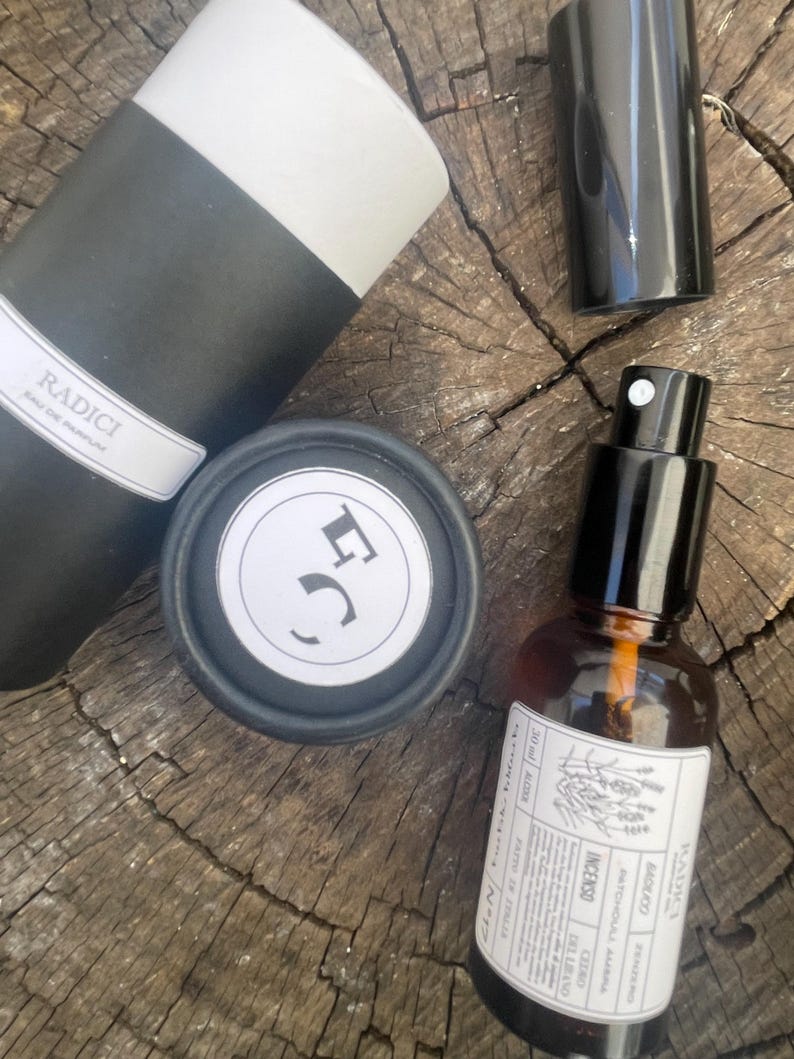 May include: A Radici Eau de Parfum set including a black and white cylindrical container, a small black cap, and an amber glass spray bottle with a white label featuring the text Radici, Patchouli, Ambra, Cedro, Incenso, and 30 ml.