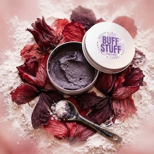 Peut inclure: Un pot en argent ouvert de gommage pour le visage "Buff Stuff" de couleur violet foncé, à côté d'une petite cuillère en céramique contenant une portion du gommage. Le pot est entouré de pétales de fleurs rouges et violettes et de poudre blanche.