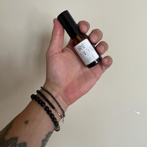 May include: A small amber glass spray bottle with a black cap and a white rectangular label featuring the text RADICI, botanical illustrations, and product details. The bottle is held upright, showcasing its compact size and dark glass construction.