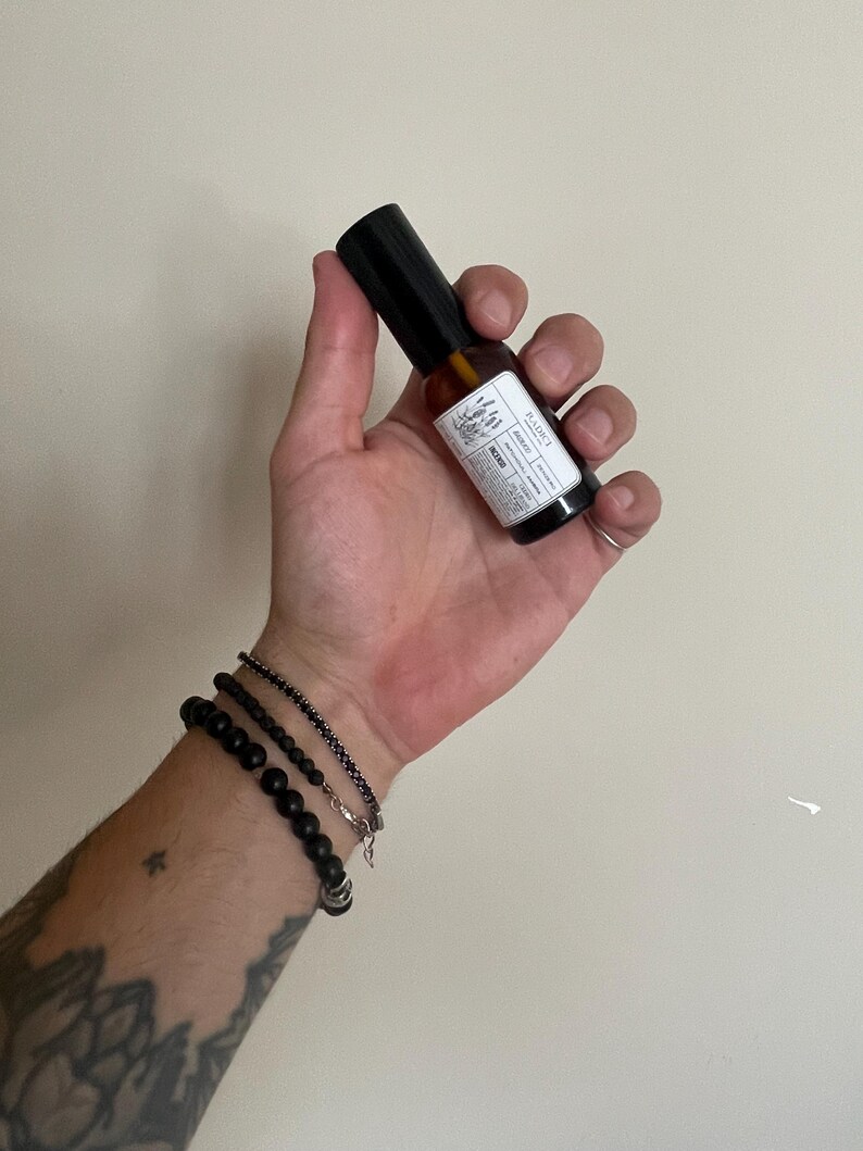May include: A small amber glass spray bottle with a black cap and a white rectangular label featuring the text RADICI, botanical illustrations, and product details. The bottle is held upright, showcasing its compact size and dark glass construction.