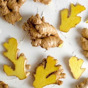 May include: Multiple pieces of fresh ginger root arranged in a scattered pattern. The ginger features light tan, papery skin with knobby, irregular shapes. Several pieces are sliced open, revealing a vibrant, bright yellow, fibrous interior flesh.