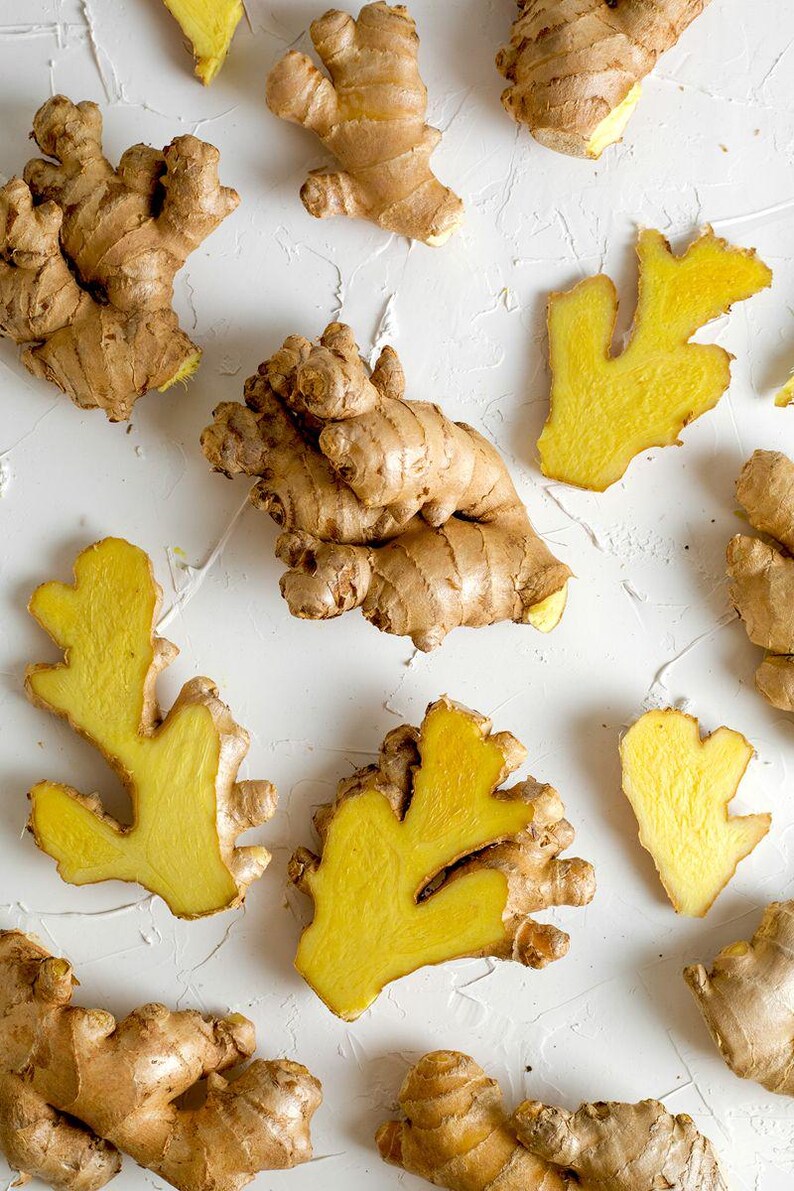 May include: Multiple pieces of fresh ginger root arranged in a scattered pattern. The ginger features light tan, papery skin with knobby, irregular shapes. Several pieces are sliced open, revealing a vibrant, bright yellow, fibrous interior flesh.