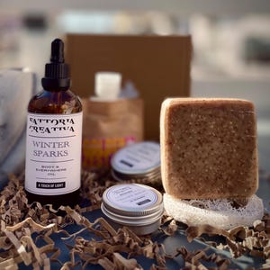 Può includere: Una scatola regalo piena di prodotti per il bagno e il corpo. La scatola contiene una bottiglia di vetro marrone con un contagocce etichettato "Fattoria Creativa Winter Sparks Body & Everywhere Oil A Touch of Light", una piccola scatola d'argento etichettata "Fattoria Creativa Walnut Balm", una saponetta marrone e una spugna di luffa bianca.