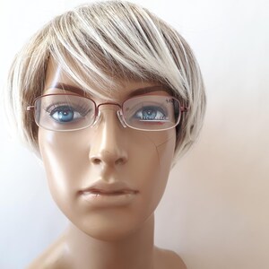 May include: A pair of rectangular eyeglasses with a thin, dark red metal frame. The lenses are clear and feature the brand name Eschenbach printed in white on the right lens. The glasses are displayed on a mannequin head with short blonde hair.