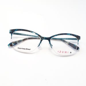 Könnte beinhalten: Brille mit einem türkis-grau gemusterten Rahmen. Die Gläser sind halb umrandet und tragen den Aufdruck "Stainless Steel" und "IZUMI". Der Rahmen ist ein dünnes Metalldesign.
