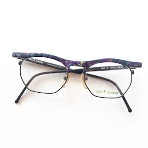 Könnte beinhalten: Vintage Brille mit einem schwarzen Metallrahmen und einer einzigartigen marmorierten Augenbrauenlinie aus lila und blauem Acetat. Die Gläser sind klar, und der Steg hat eine goldfarbene Oberfläche. Die Brille ist mit "Best Company" und "Hand Made Italy" gekennzeichnet.