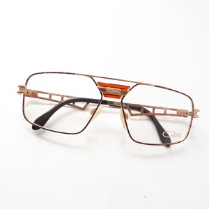 May include: Gold-coloured eyeglasses with a rectangular frame and a double bridge. The glasses have clear lenses and dark temples. An orange accent is visible on the bridge. The brand name is printed on the lens.