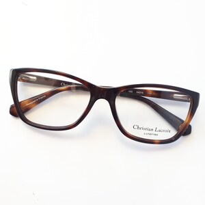 May include: Brown tortoiseshell eyeglasses with a cat-eye shape. The frames have a dark brown color with lighter brown accents. The brand name "Christian Lacroix" is printed on the right temple.