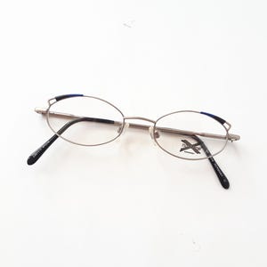Könnte beinhalten: Ovale Brille mit silbernem Metallrahmen und schwarzen Bügeln. Die Oberseite des Rahmens hat einen kleinen blauen Akzent. Die Gläser sind klar. Die Brille hat ein kleines Etikett mit Text auf dem rechten Glas.
