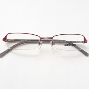 Könnte beinhalten: Rote Metallgestellbrille mit klaren Gläsern. Der Markenname "MOSCHINO" ist auf dem rechten Glas aufgedruckt.