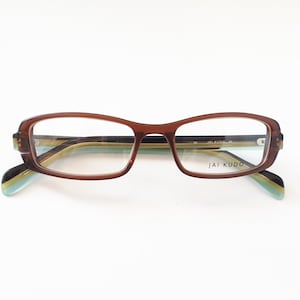 Könnte beinhalten: Braune rechteckige Brille mit einem schwarz-grünen gestreiften Bügel. Der Markenname "JAI KUDO" ist auf dem rechten Bügel aufgedruckt.