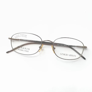 May include: Titanio nickel-free eyeglasses with a Giorgio Armani logo. The oval frames are a dark brown color. The glasses have a clip-on sunglasses feature.