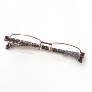 Puede incluir: Gafas con montura de metal bronce y diseño estampado en las patillas. Las lentes son transparentes y las gafas tienen un diseño de media montura. La marca "FACE FACE" es visible en la lente.