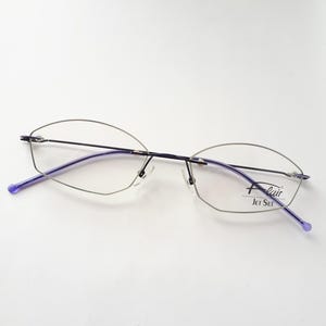 Könnte beinhalten: Eine silberfarbene, randlose Brille mit violetten Bügeln. Die Brille hat eine rechteckige Form und der Markenname "Flair Jet Set" ist auf dem rechten Bügel aufgedruckt.