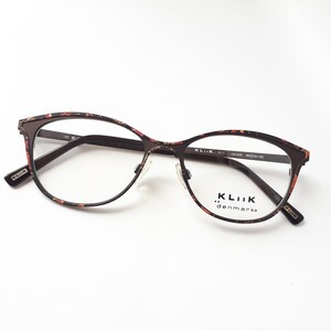 May include: Brown and pink patterned metal eyeglass frames with clear lenses. The frames have a cat-eye shape and the brand name "KLIIK... denmark" is printed on the right lens.