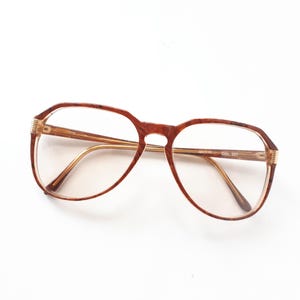 May include: Brown-framed eyeglasses with a patterned design. The glasses have a rectangular shape with rounded edges and gold-coloured arms. The lenses are clear, and the frame has a textured appearance. The glasses are set against a white background.