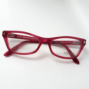 Może przedstawiać: Okulary w czerwonych oprawkach z tworzywa sztucznego z napisem "1-2/12 TOM FORD MADE IN ITALY" na prawej zauszniku i "TOM FORD" na prawej soczewce.