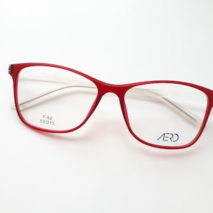Könnte beinhalten: Brille mit rotem Rahmen und rechteckiger Form. Die Brille hat klare Gläser und der Markenname "AERO" ist auf dem unteren rechten Glas aufgedruckt. Die Rahmengröße ist F-62 53015.