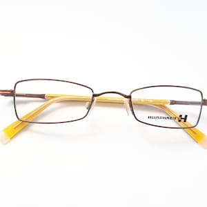 Könnte beinhalten: Braune Metallbrille mit gelben Bügeln und einem schwarzen Humphrey's H-Logo auf der rechten Linse.
