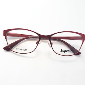 Puede incluir: Gafas de metal con montura roja y lentes transparentes. El nombre de la marca "Superflex Titanium" está impreso en la patilla derecha.