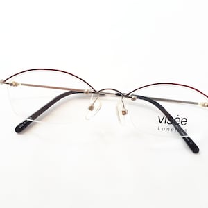 Könnte beinhalten: Silberne randlose Brille mit einem burgunderfarbenen Akzent auf dem oberen Rahmen. Der Markenname "visée Lunettes" ist auf dem rechten Bügel aufgedruckt.