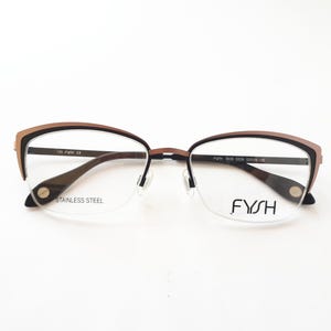 Könnte beinhalten: Braune und schwarze Cat-Eye-Brille mit klaren Gläsern. Die Brille hat einen Metallrahmen und die Worte "STAINLESS STEEL" und "FYSH" sind auf den Bügeln aufgedruckt.