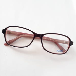 Könnte beinhalten: Brille mit einem dunkelburgunderfarbenen Rahmen und hellrosa Bügeln. Die Gläser sind klar, und der Markenname "JOOP" ist auf dem unteren rechten Glas aufgedruckt. Die Brille befindet sich vor einem weißen Hintergrund.