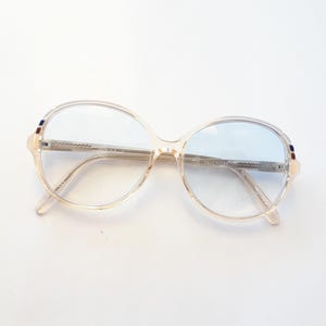 May include: Vintage oversized spectacles with a light beige frame and blue-tinted lenses. The glasses have a rounded shape and thin metal arms. Small accents of red, white, and blue are on the frame.
