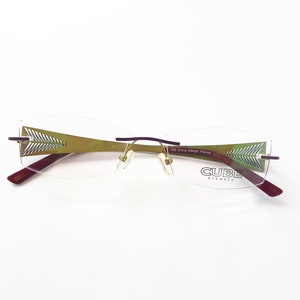 Könnte beinhalten: Eine Brille ohne Rahmen mit goldenem Gestell und violetten Bügeln. Die Gläser sind klar und die Bügel haben ein dekoratives Muster. Der Markenname "CUBE EYEWEAR" ist auf dem rechten Glas aufgedruckt.