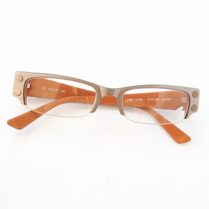 May include: A pair of brown and silver eyeglasses with clear lenses. The frame has a subtle cat-eye shape and features small metal accents on the temples. The text "CK 31018 140" is printed on the right temple and "Ref. Lumina 8118-03E By AMA" is printed on the left temple.