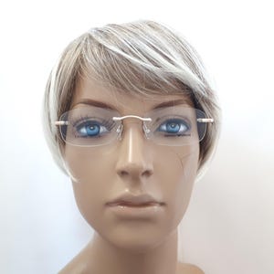 May include: Rimless eyeglasses with a gold-colored bridge and arms. The lenses are clear, and the brand name "Emporio Armani" is visible on the lens. The glasses are on a mannequin with blue eyes and a short, light-colored wig.
