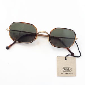 May include: Vintage sunglasses with a rectangular shape and a tortoiseshell pattern frame. The lenses are dark green, and the arms are brown. A Safilo Group tag is attached.