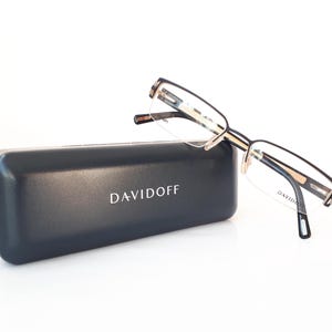 Könnte beinhalten: Schwarze rechteckige Brillenbox mit dem in Weiß gedruckten Wort "DAVIDOFF". Die Box ist geschlossen und liegt neben einer Brille mit schwarzem und goldenem Rahmen.