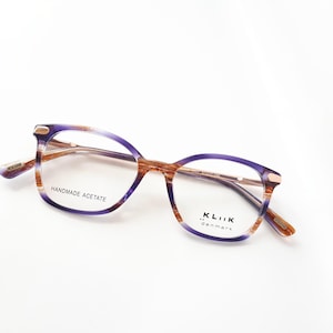 Könnte beinhalten: Lila und braune Acetat-Brille mit einer goldenen Metallbrücke und Bügeln. Die Gläser sind klar und die Worte "HANDMADE ACETATE" und "KLIK DENMARK" sind auf den Rahmen gedruckt.