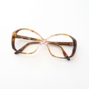 Könnte beinhalten: Vintage Brille mit braunem und beigefarbenem Schildpattmuster. Die Fassungen haben eine große, abgerundete Form mit klaren Gläsern. Die Bügel sind hellbraun. Die Brille liegt vor einem weißen Hintergrund.